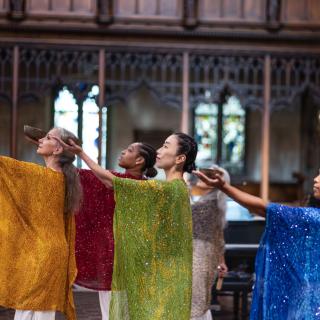 Omega Dance Company performers in glittering smocks each hold bowls and extend their arms toward the sky. They perform in a neo-Gothic church.