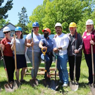 At a groundbreaking ceremony, a line of eight smiling people wearing construction hats stand with shovels on a vacant lot that will become an affordable housing community.