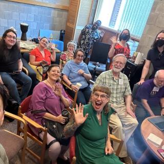 A smiling group of people sit in a room together and pose for a photo to mark their getting together to experience the online Unitarian Universalist Association General Assembly 2024.