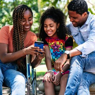 Three people -- two adults and a child -- sit together at a park on a sunny day and smile as they look at something on a smartphone.