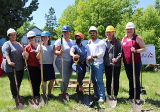 At a groundbreaking ceremony, a line of eight smiling people wearing construction hats stand with shovels on a vacant lot that will become an affordable housing community.