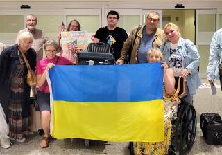 A group of people in an airport pose together while some of them hold up a large Ukrainian flag. 