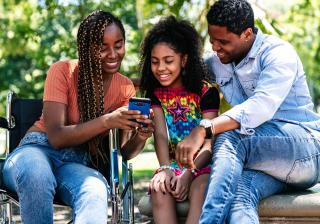 Three people -- two adults and a child -- sit together at a park on a sunny day and smile as they look at something on a smartphone.