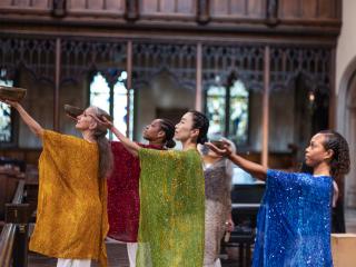 Omega Dance Company performers in glittering smocks each hold bowls and extend their arms toward the sky. They perform in a neo-Gothic church.