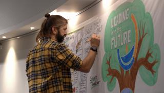 A person sketches a mural during a conference that happened in Prague. The mural has a big tree with the words "Leading into the Future" and the muralist is adding images that depict collaborative conversations happening at the conference.
