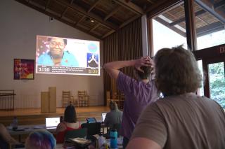 Facing a large projector screen, a group of people react to a live closing speech given during the 2024 Unitarian Universalist Association General Assembly.