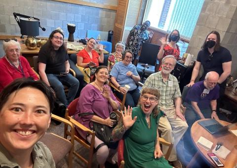 A smiling group of people sit in a room together and pose for a photo to mark their getting together to experience the online Unitarian Universalist Association General Assembly 2024.